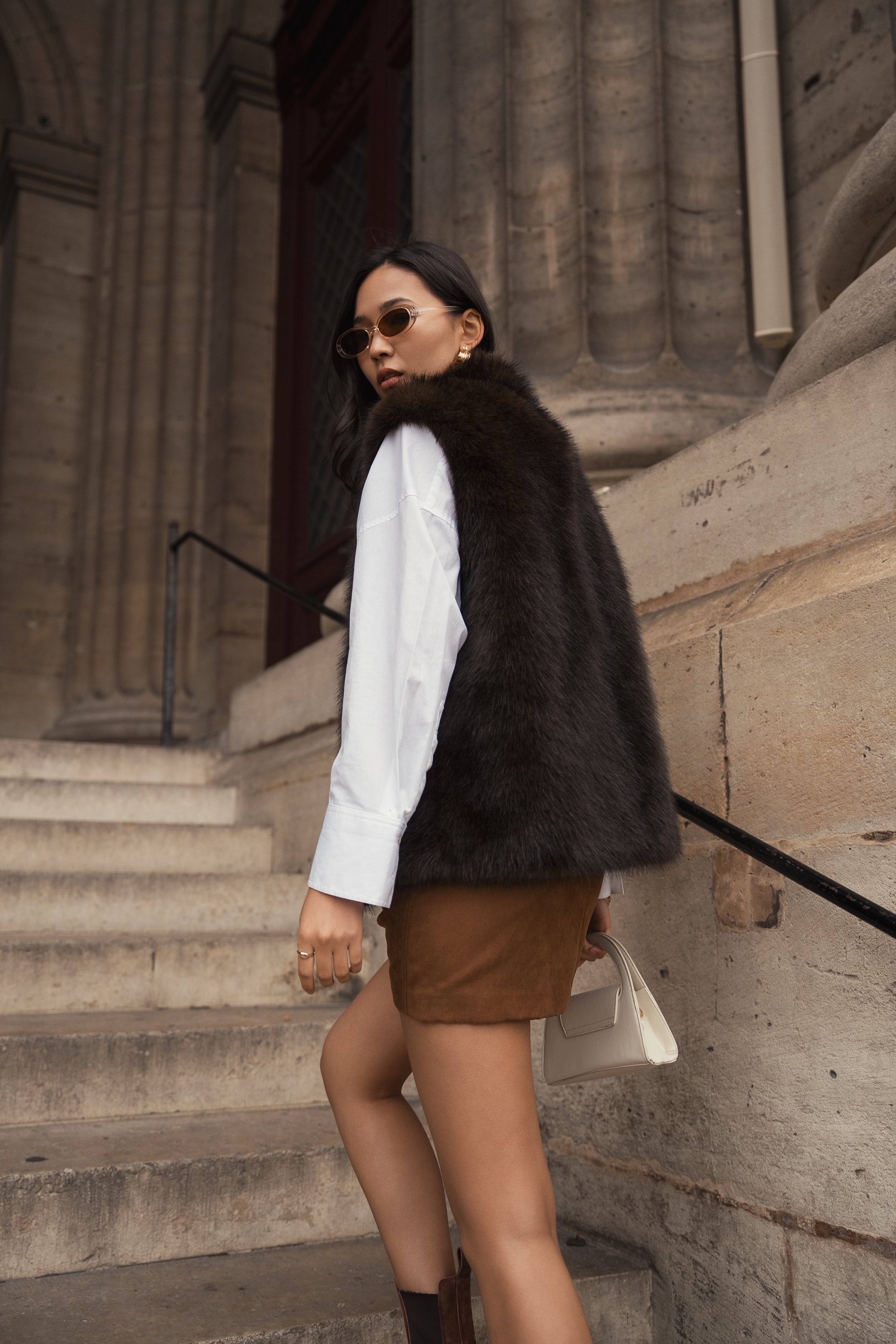 Femme portant le gilet sans manches CAMELIA en fausse fourrure chocolat Escandelle – look chic d’extérieur avec chemise blanche et jupe camel
#couleur_choco