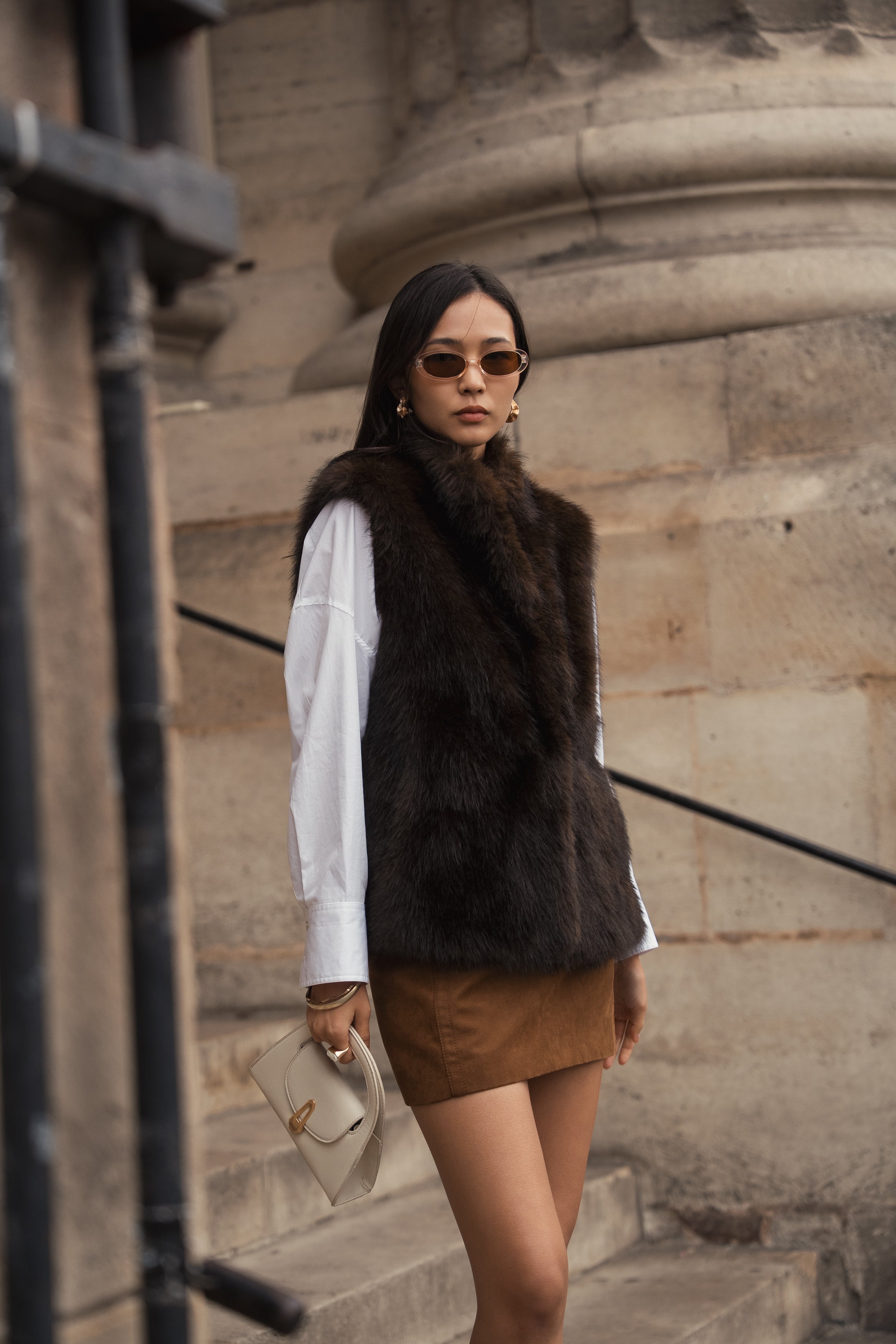 Femme portant le gilet sans manches CAMELIA en fausse fourrure chocolat Escandelle – look chic d’extérieur avec chemise blanche et jupe camel
#couleur_choco