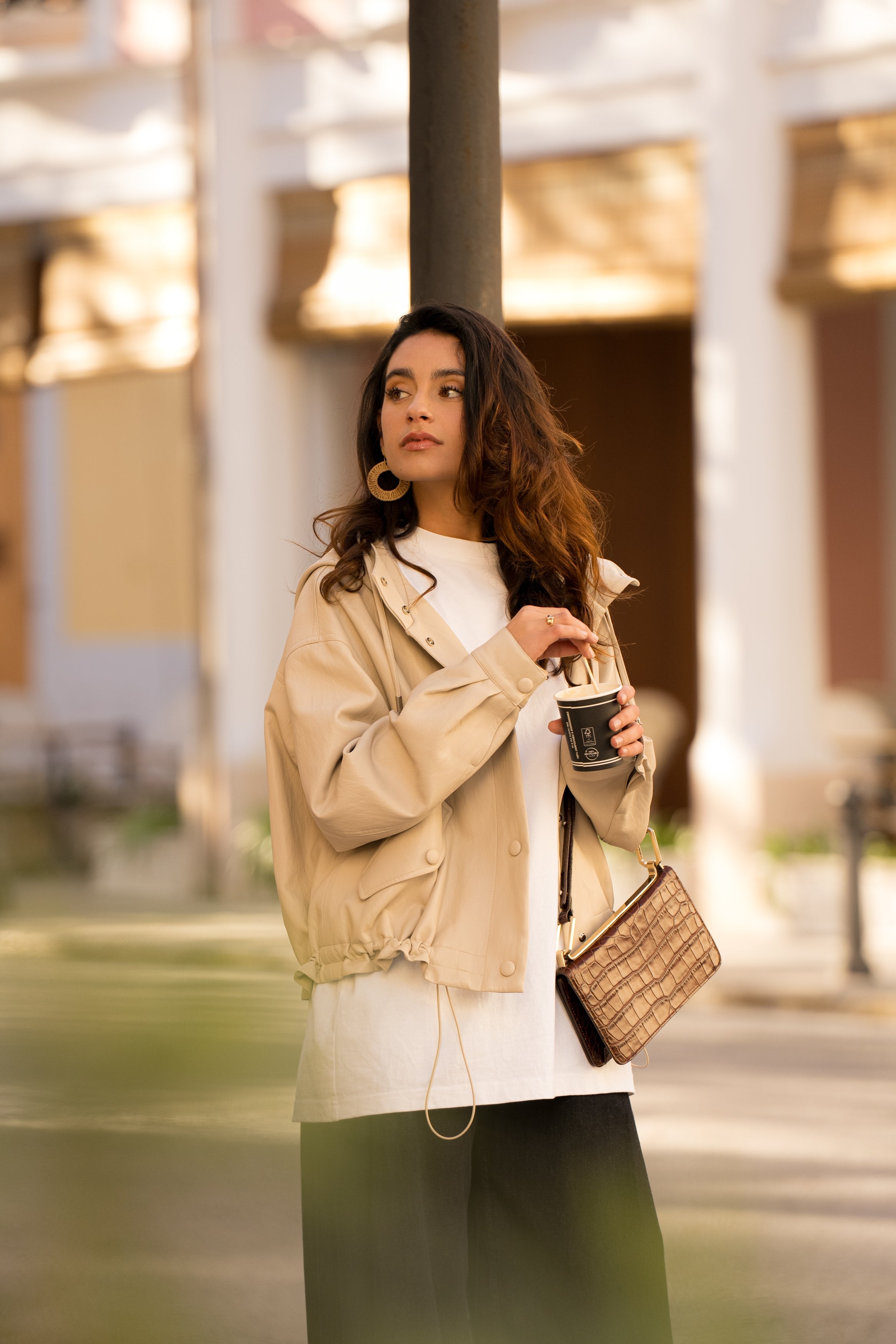 femme portant une tenue confortable chic pour les vacances ou le quotidien avec un blouson en similicuir, fermeture à boutons pression, manches larges refermées par un bouton au poignet, capuche à cordons, poches à rabats et bas ajustable avec élastique, côté
#couleur_beige