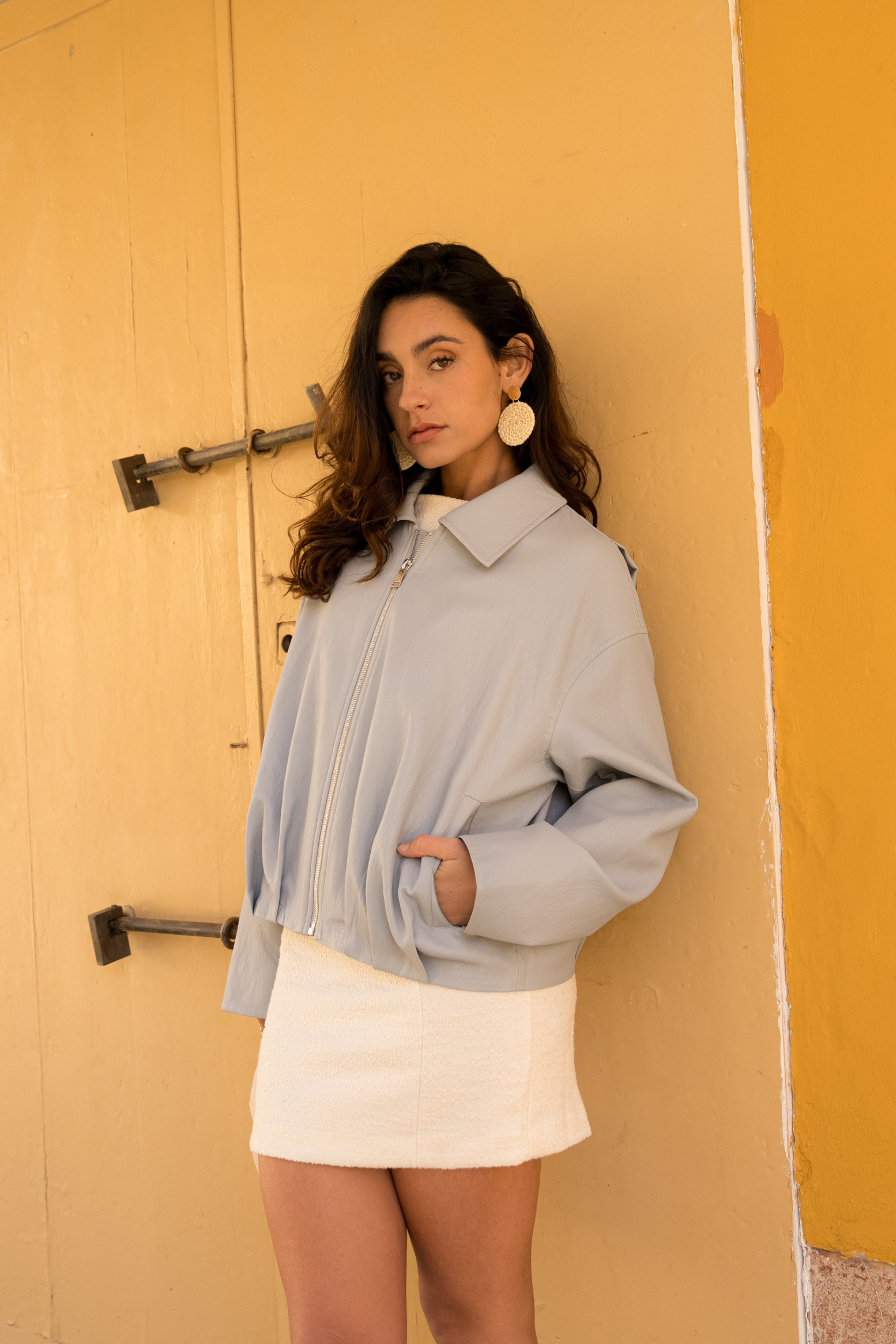 blouson en similicuir à col français, coupe droite, bas plissé devant dos, fermeture à glissière métallique, style streetwear féminin unisexe, devant, femme qui porte la veste serina avec une main dans la poche raglans côté
#couleur_ciel