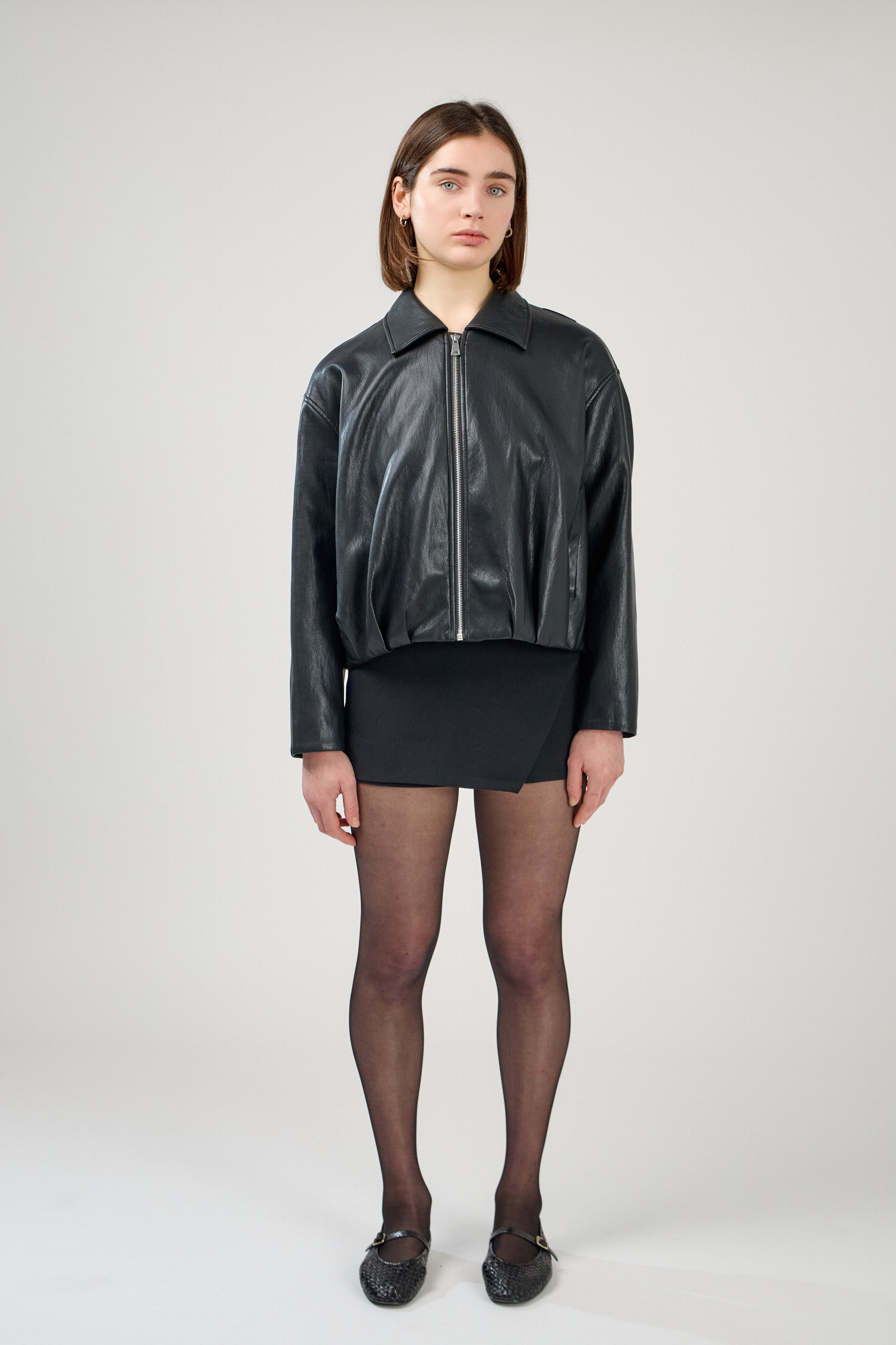 blouson en similicuir à col français, coupe droite, bas plissé devant dos, fermeture à glissière métallique, style streetwear féminin unisexe, devant, porté avec une jupe courte noire
#couleur_noir