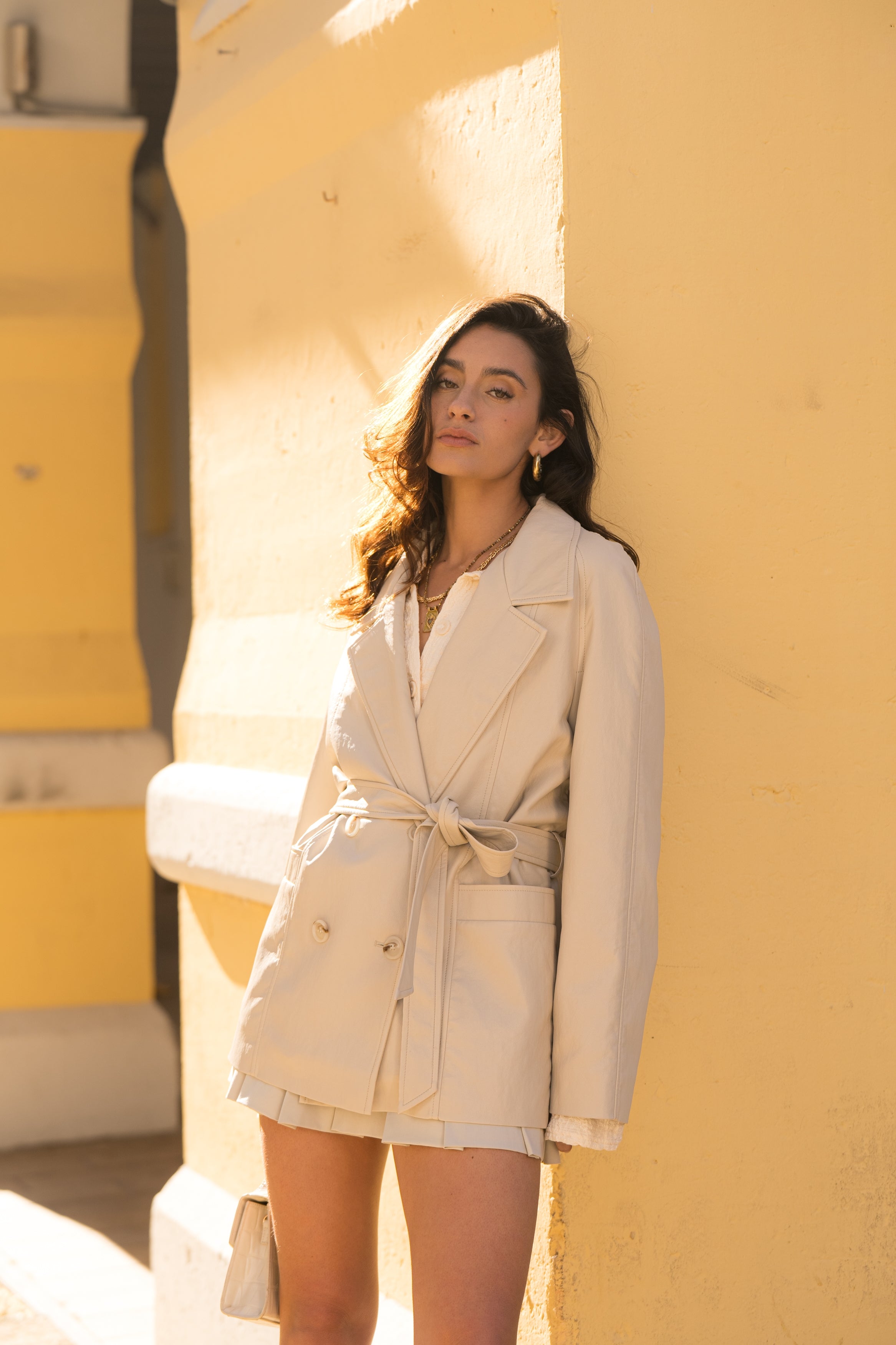 Femme portant veste ovania beige clair en similicuir, veste de tailleur col à revers, découpes verticales, fermeture à boutons, ceinture et passants, devant
#couleur_beige