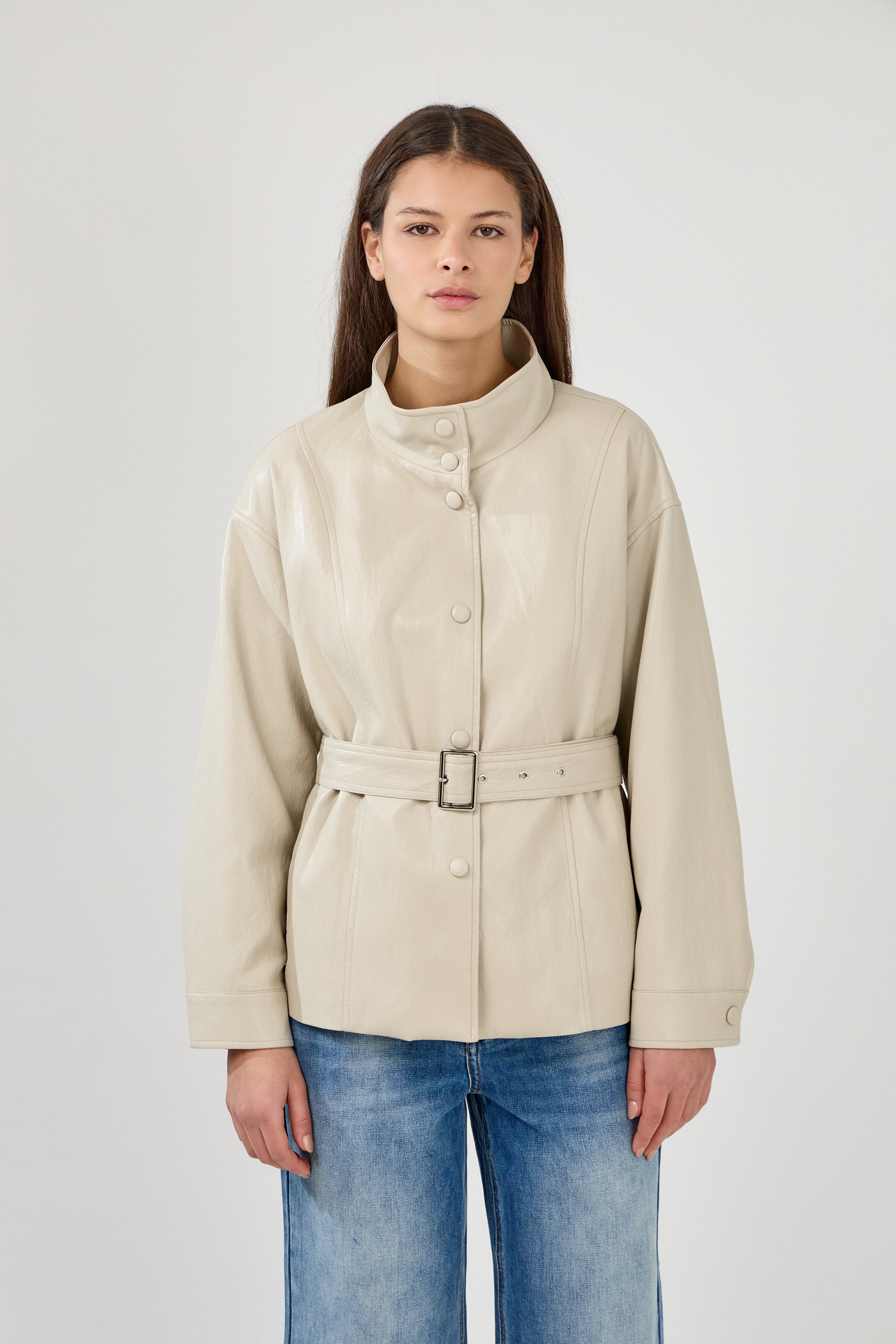 femme portant une veste en similicuir avec une ceinture à boucle métallique et fermeture à boutons pressions. Manches longues et boutons poignets. Découpe verticales devant, col montant, devant
#couleur_beige