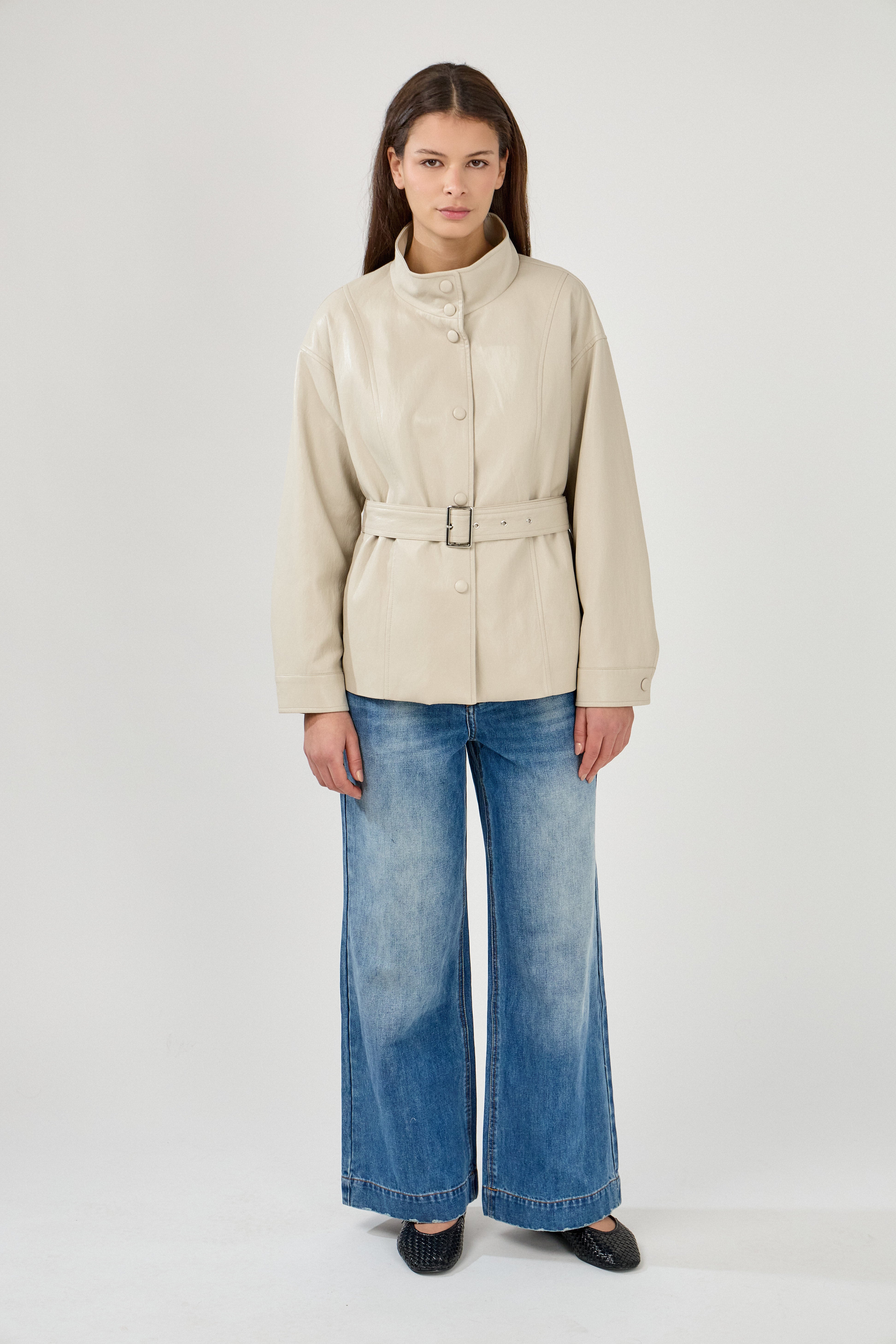 femme portant une veste en similicuir avec une ceinture à boucle métallique et fermeture à boutons pressions. Manches longues et boutons poignets. Découpe verticales devant, col montant, devant
#couleur_beige