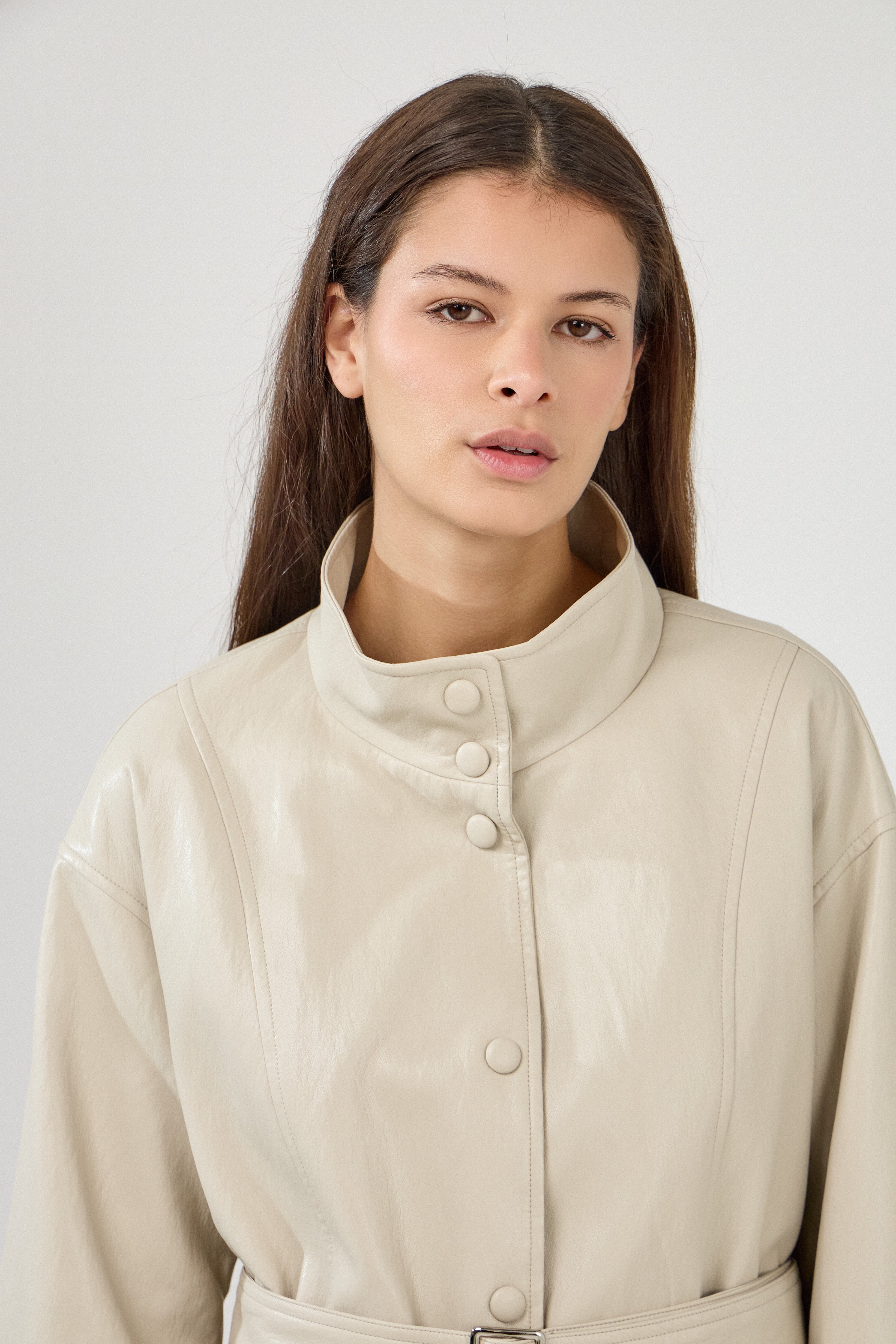 femme portant une veste en similicuir avec une ceinture à boucle métallique et fermeture à boutons pressions. Manches longues et boutons poignets. Découpe verticales devant, col montant, zoom devant
#couleur_beige