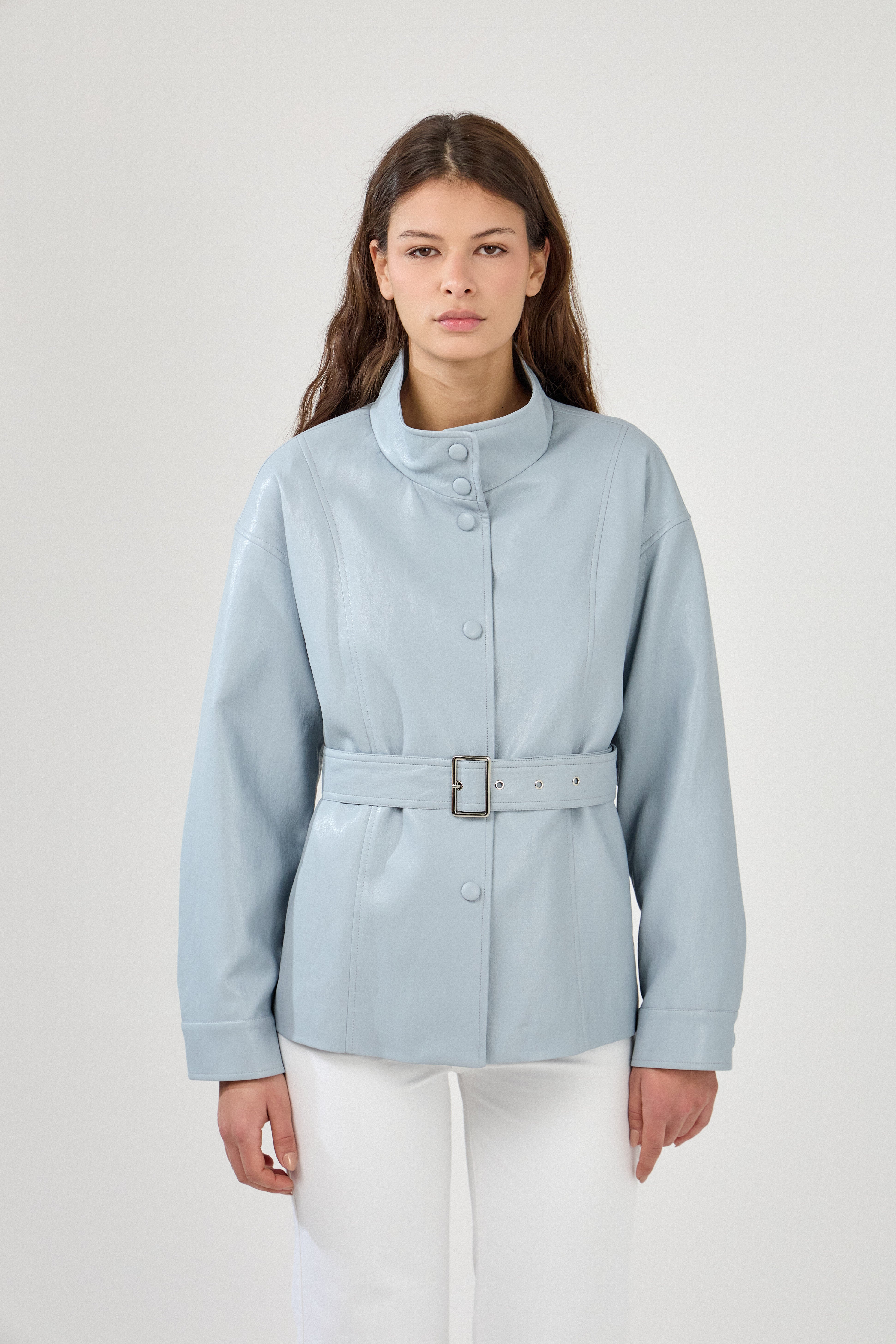 femme portant une veste en similicuir avec une ceinture à boucle métallique et fermeture à boutons pressions. Manches longues et boutons poignets. Découpe verticales devant, col montant, devant
#couleur_ciel