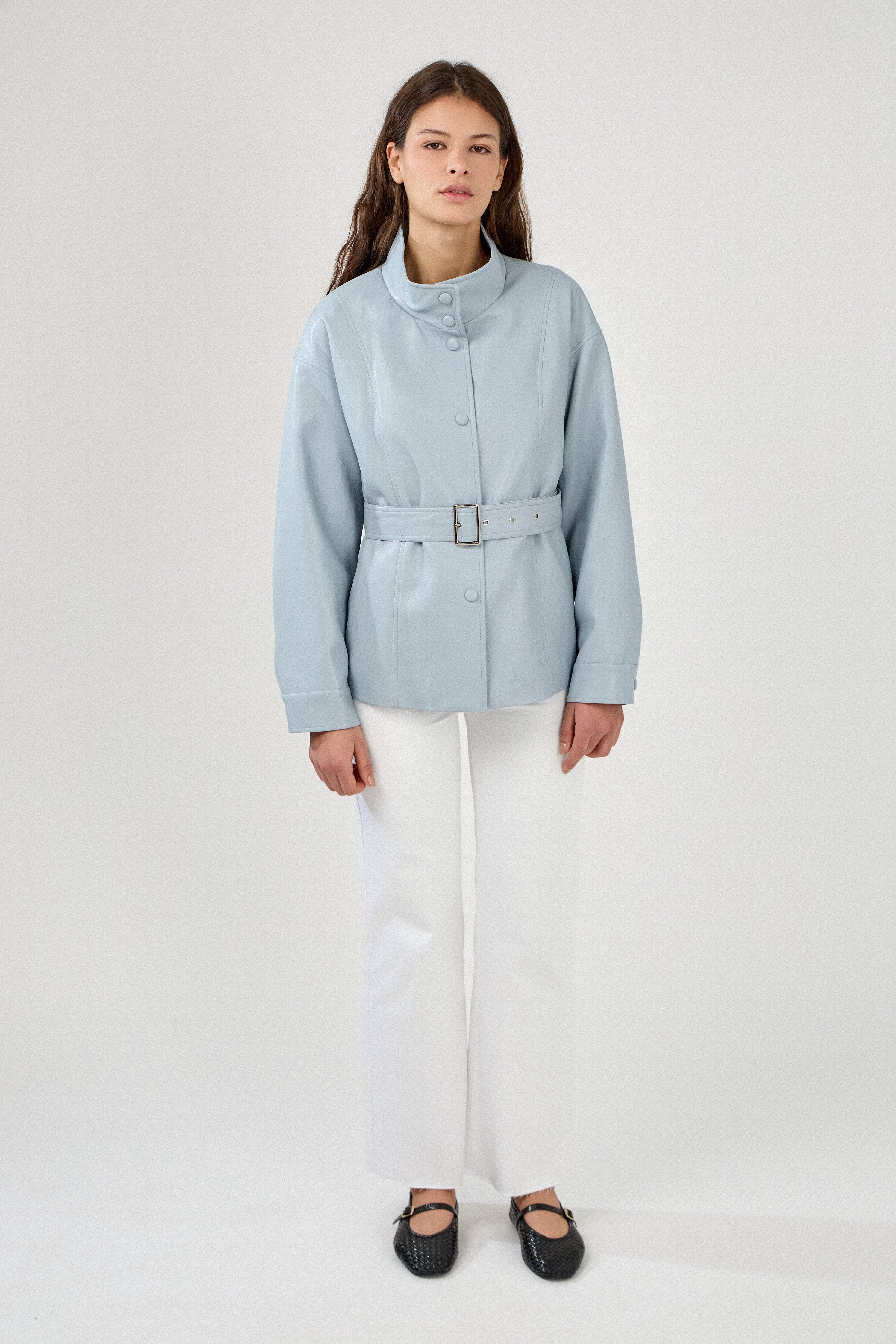 femme portant une veste en similicuir avec une ceinture à boucle métallique et fermeture à boutons pressions. Manches longues et boutons poignets. Découpe verticales devant, col montant, devant
#couleur_ciel