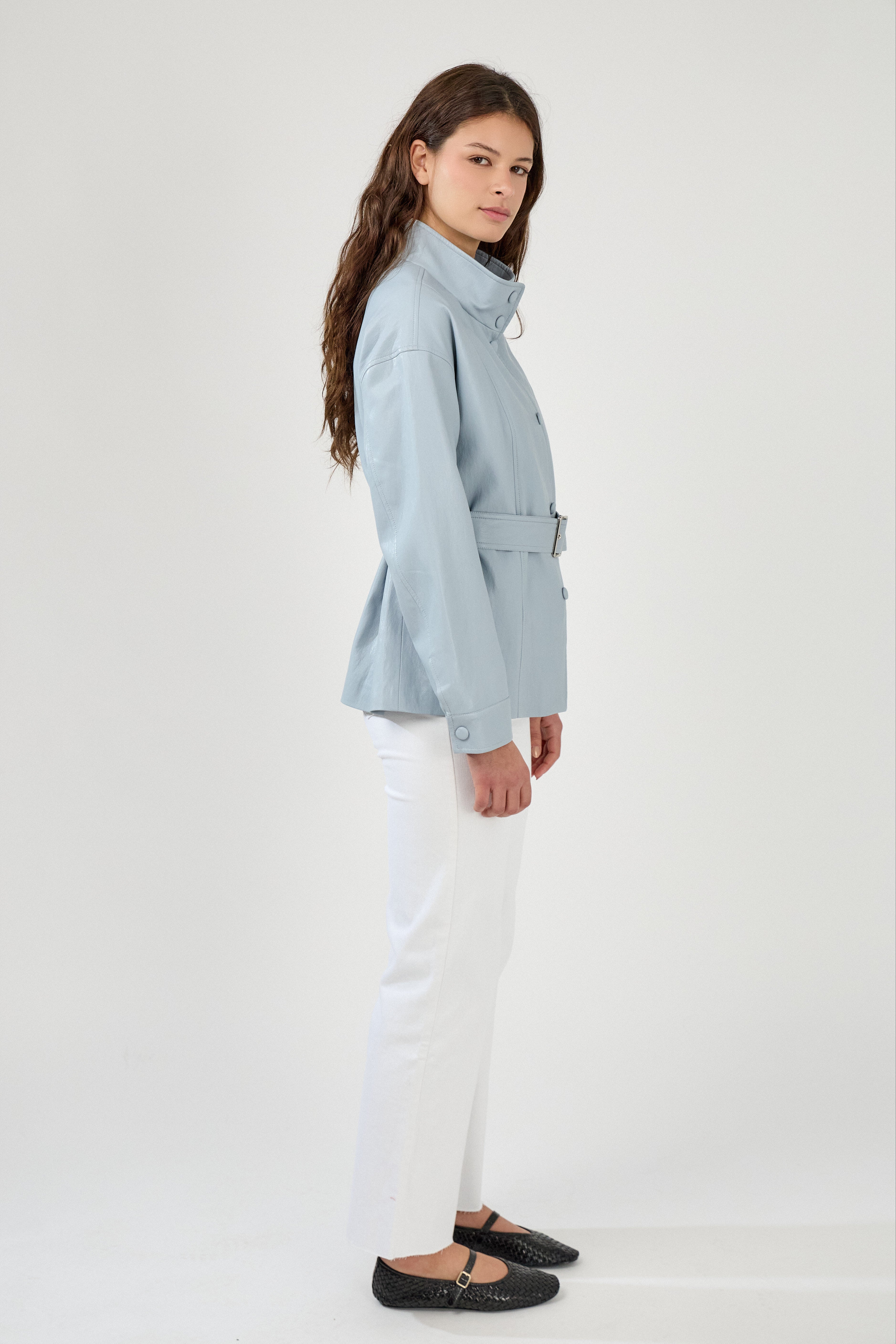 femme portant une veste en similicuir avec une ceinture à boucle métallique et fermeture à boutons pressions. Manches longues et boutons poignets. Découpe verticales devant, col montant, côté
#couleur_ciel