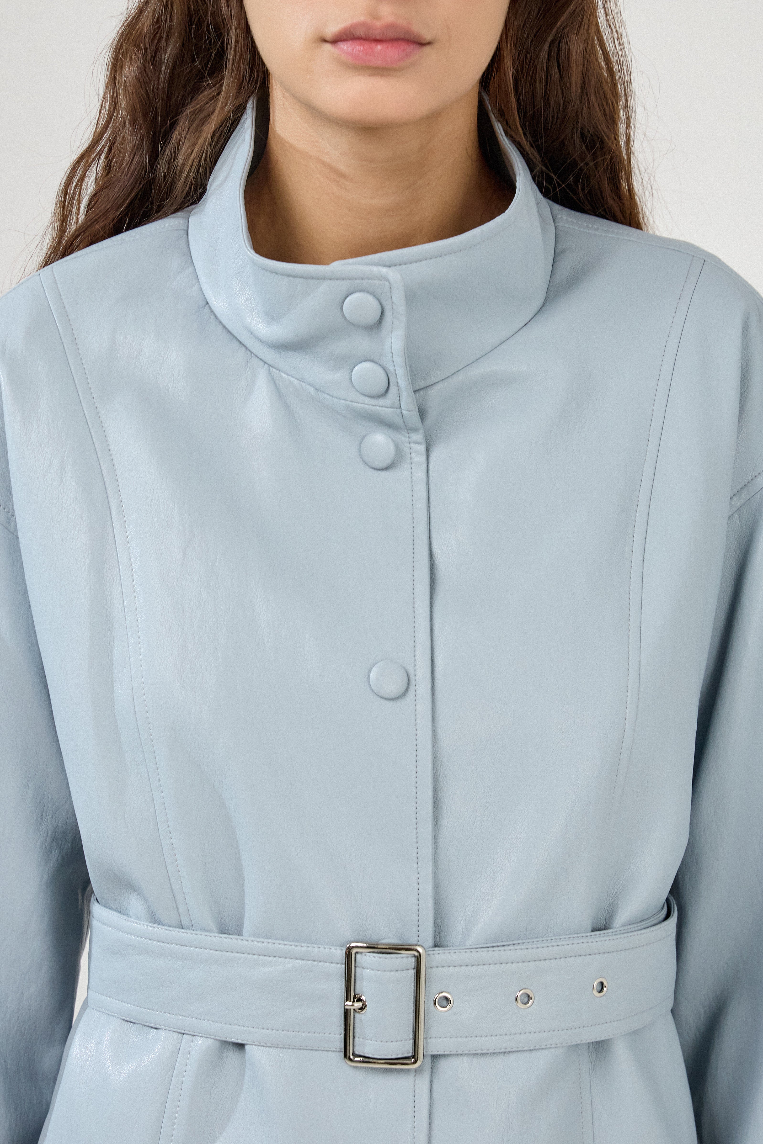 femme portant une veste en similicuir avec une ceinture à boucle métallique et fermeture à boutons pressions. Manches longues et boutons poignets. Découpe verticales devant, col montant, zoom devant
#couleur_ciel