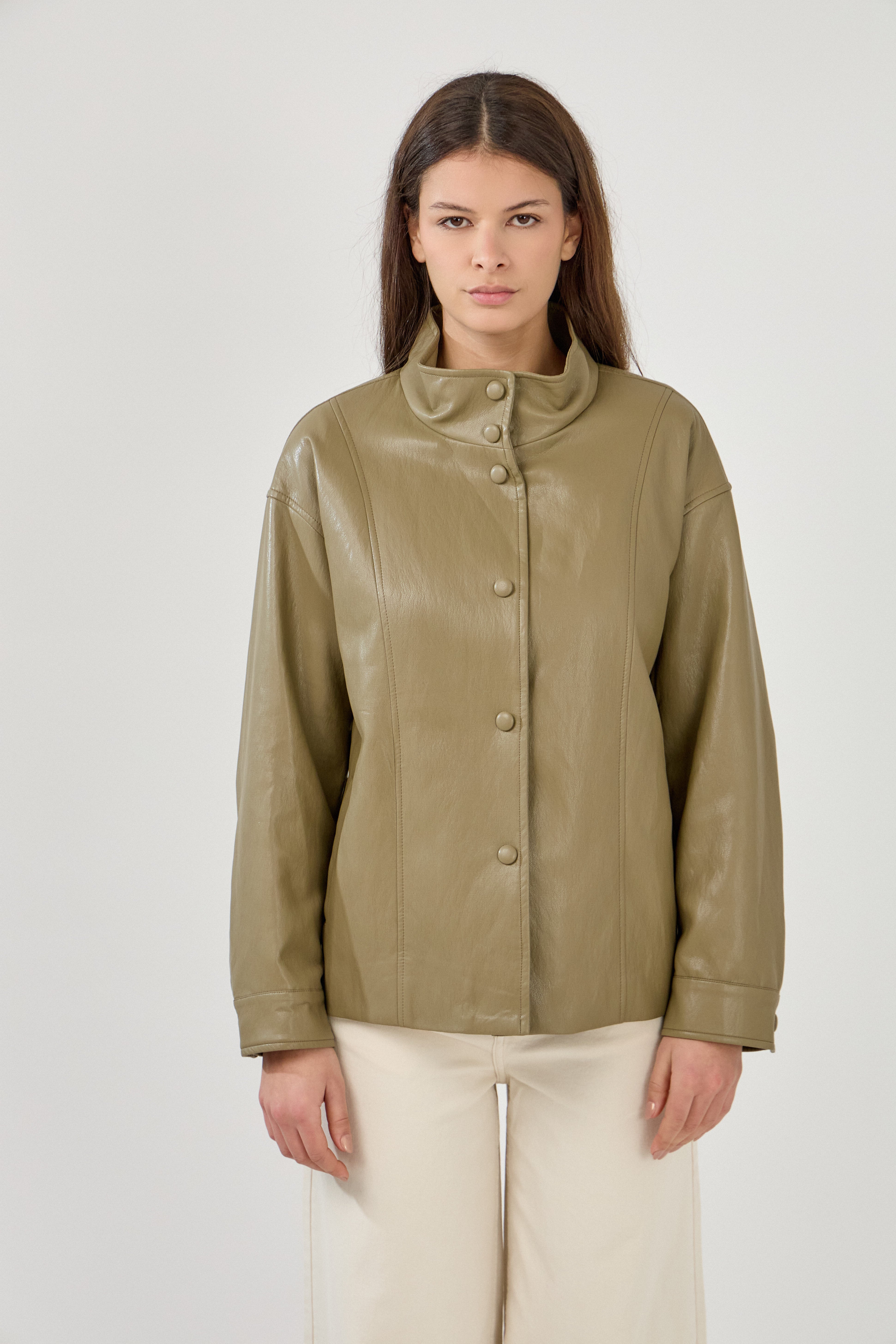 femme portant une veste en similicuir, portée sans ceinture. fermeture à boutons pressions. Manches longues et boutons poignets. Découpe verticales devant, col montant, devant
#couleur_kaki