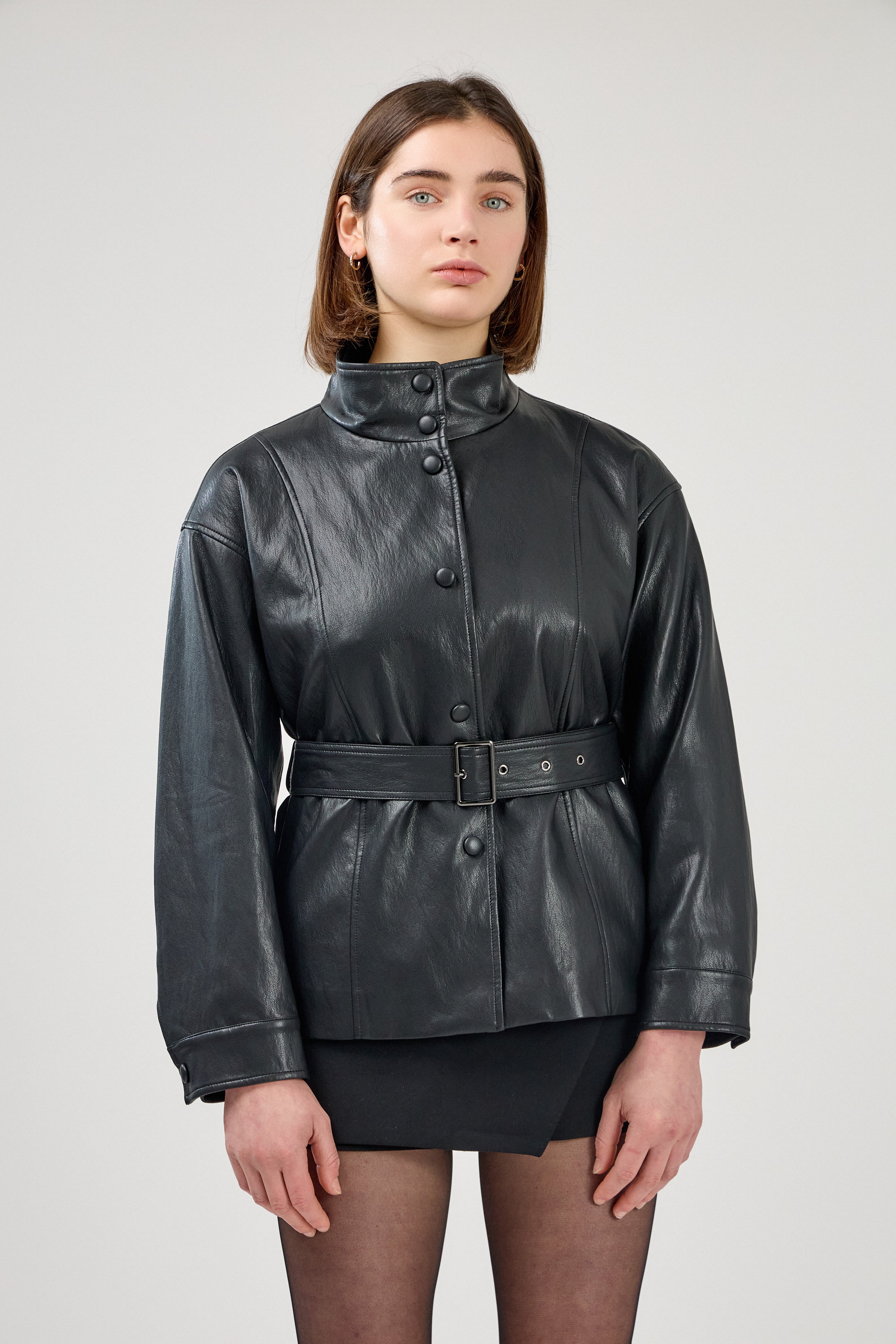 femme portant une veste en similicuir avec une ceinture à boucle métallique et fermeture à boutons pressions. Manches longues et boutons poignets. Découpe verticales devant, col montant, devant
#couleur_noir