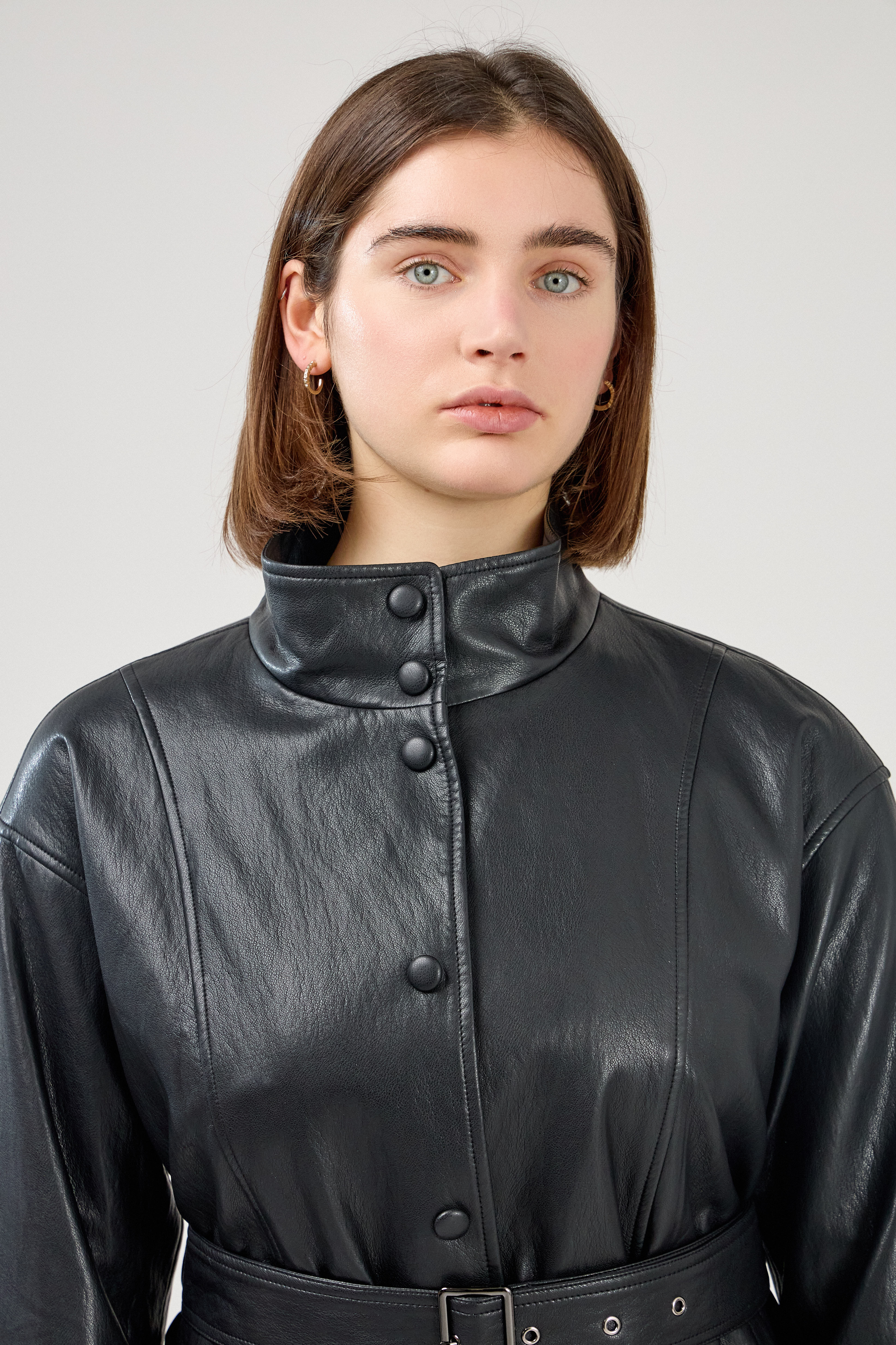 femme portant une veste en similicuir avec une ceinture à boucle métallique et fermeture à boutons pressions. Manches longues et boutons poignets. Découpe verticales devant, col montant, zoom devant
#couleur_noir