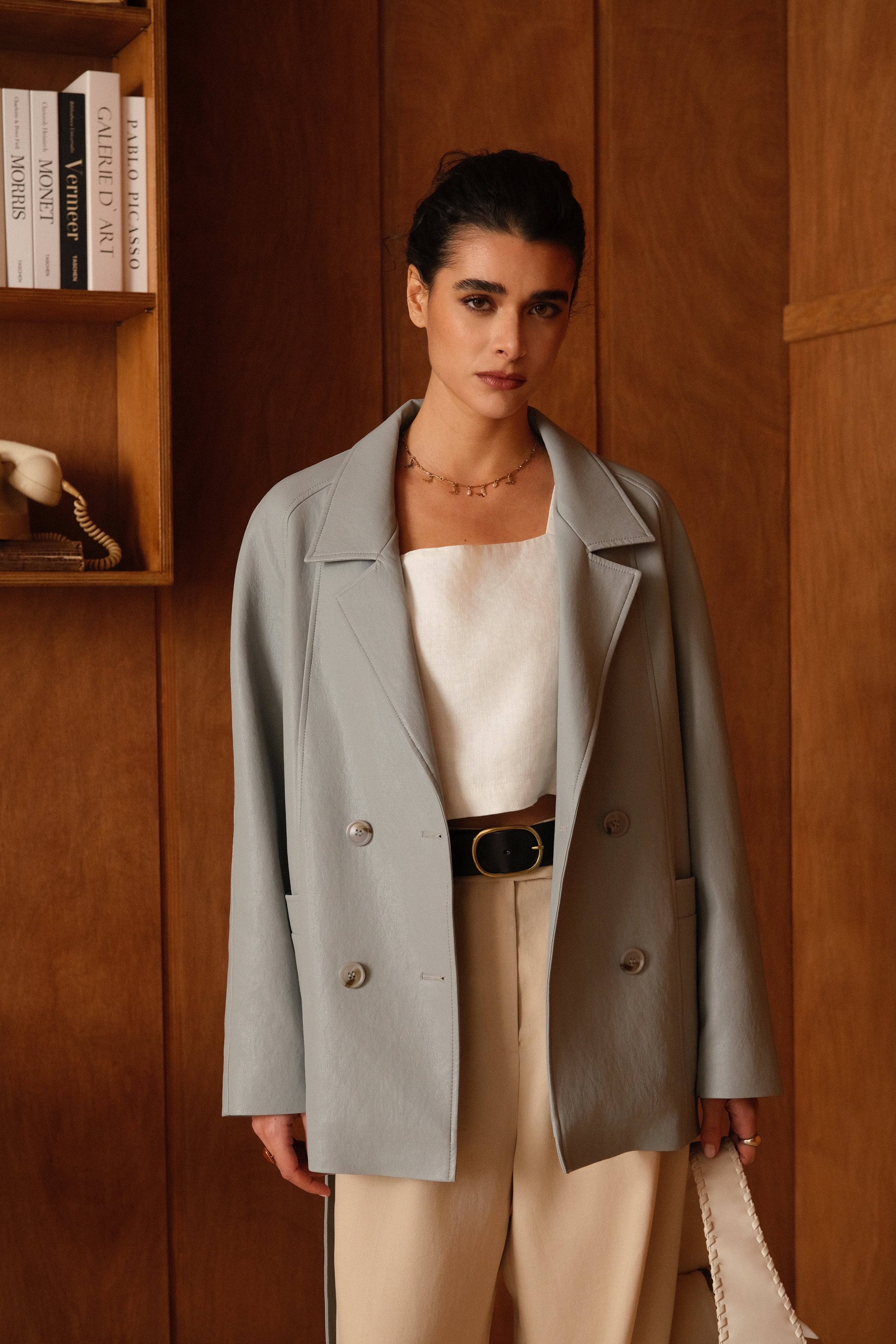 Femme portant veste ovania bleu ciel en similicuir, veste de tailleur col à revers, découpes verticales, fermeture à boutons, ceinture et passants, devant
#couleur_ciel