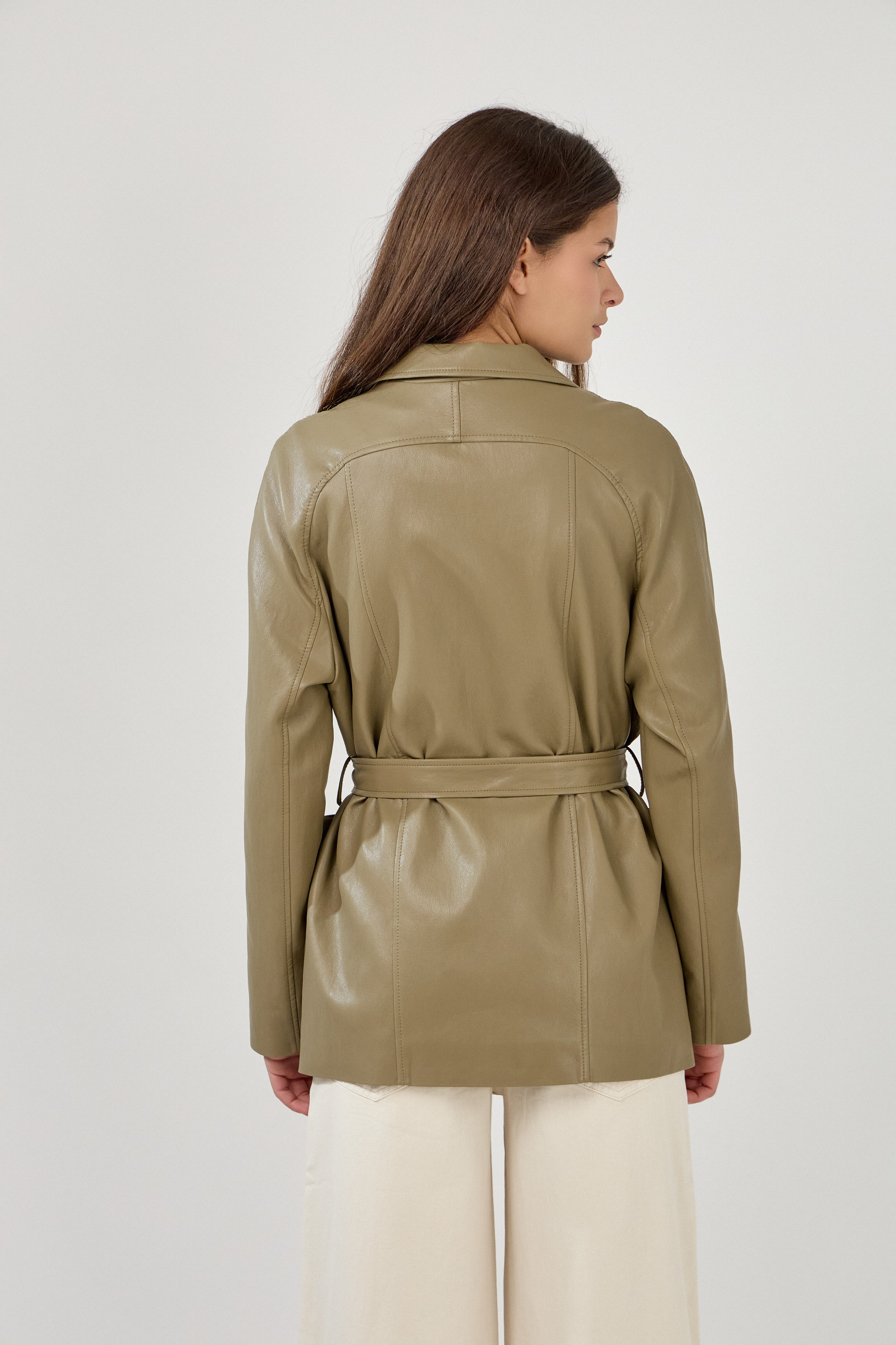 Femme portant veste ovania kaki en similicuir, veste de tailleur col à revers, découpes verticales, fermeture à boutons, ceinture et passants, dos
#couleur_kaki