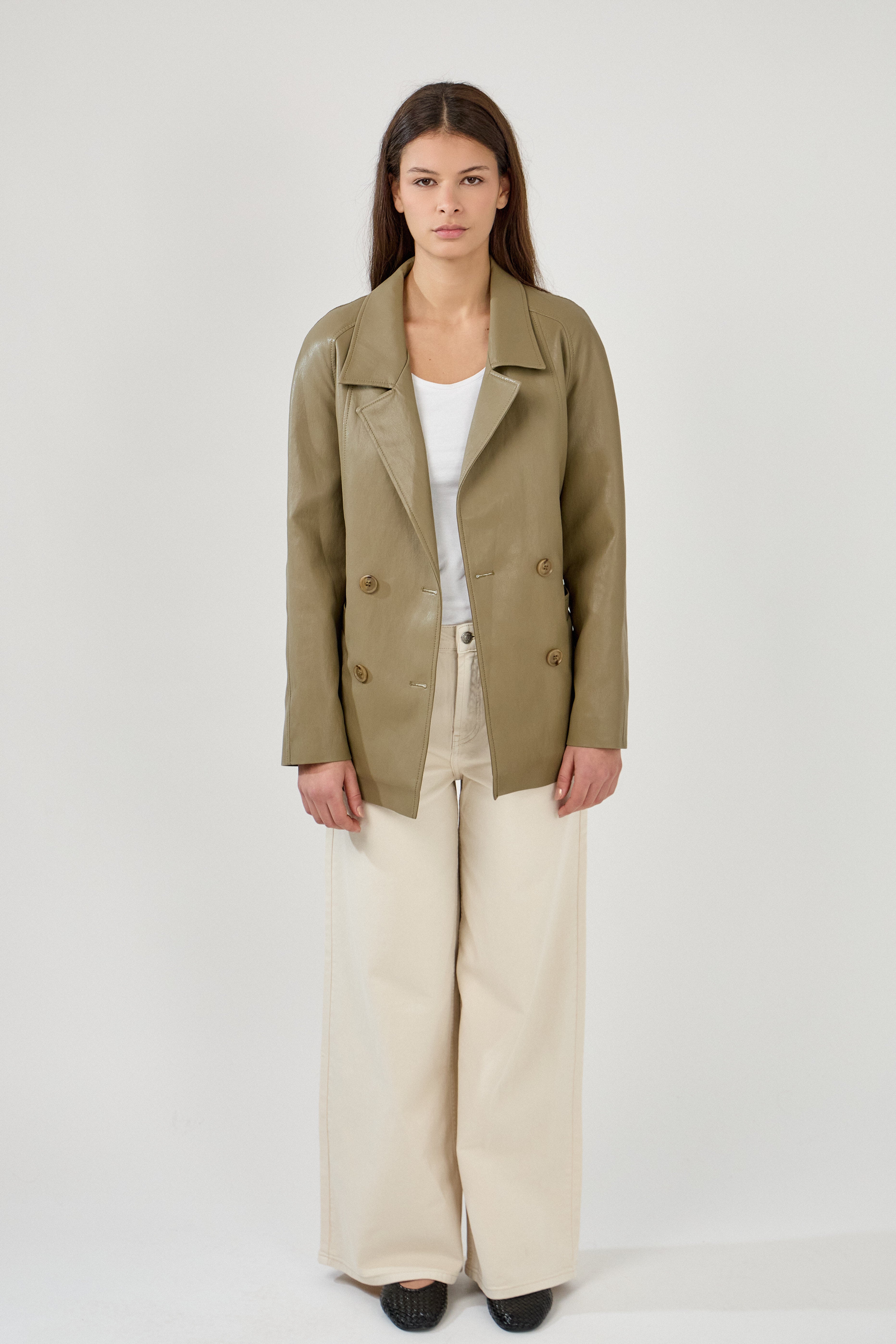 Femme portant veste ovania kaki en similicuir, veste de tailleur col à revers, découpes verticales, fermeture à boutons, ceinture et passants, devant
#couleur_kaki