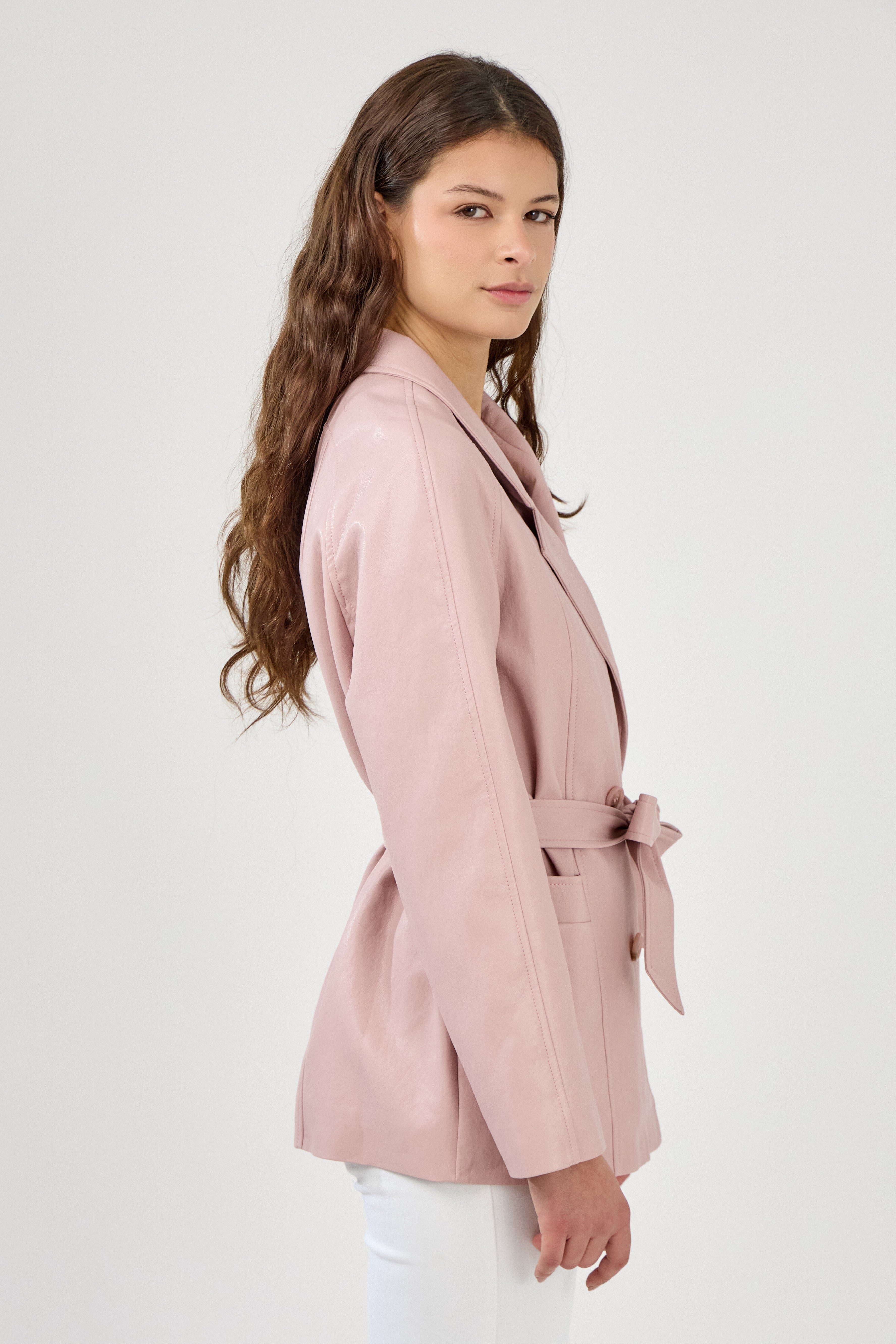 Femme portant veste ovania rose pâle en similicuir, veste de tailleur col à revers, découpes verticales, fermeture à boutons, ceinture et passants, côté
#couleur_rose