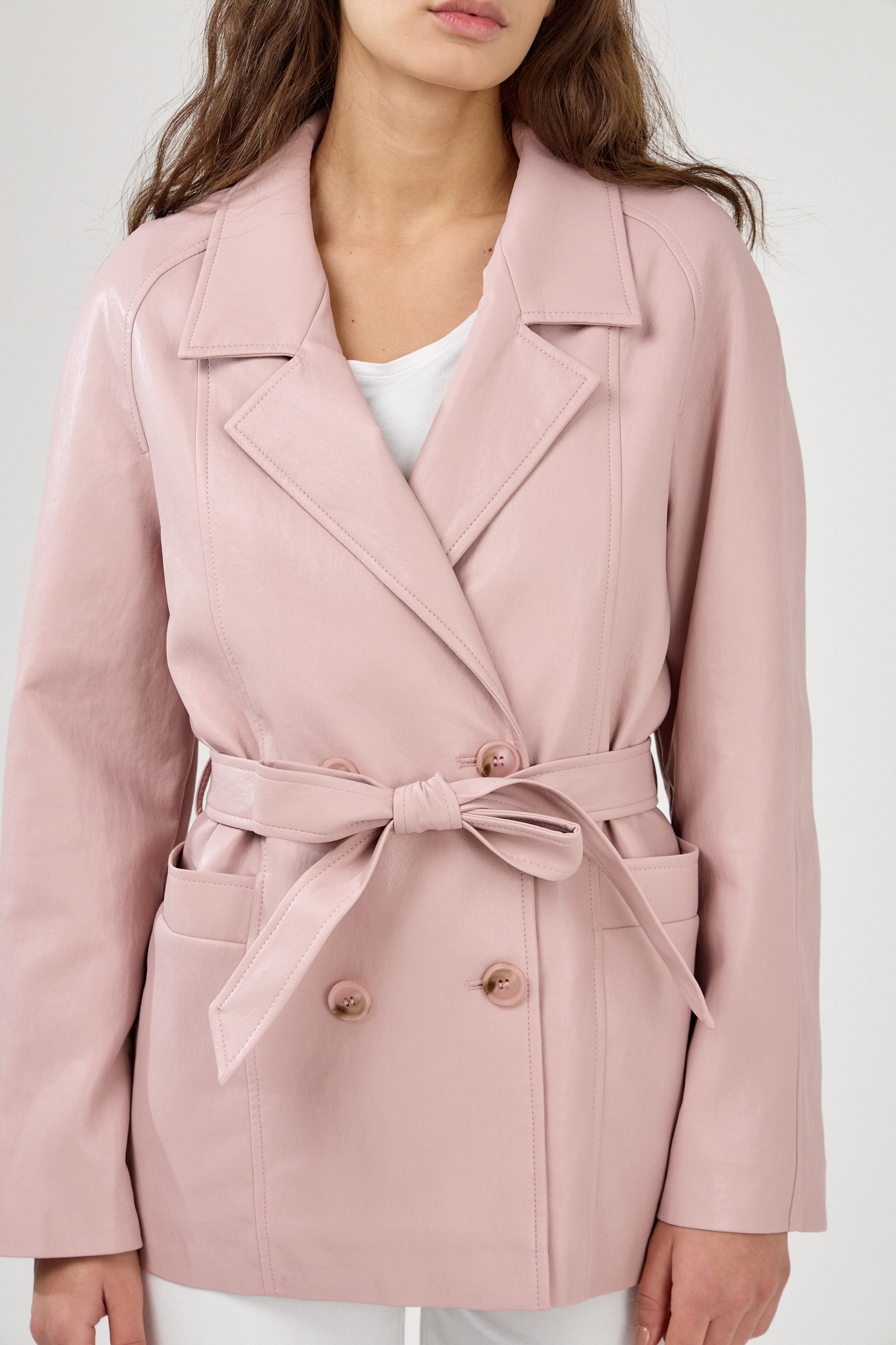 Femme portant veste ovania rose pâle en similicuir, veste de tailleur col à revers, découpes verticales, fermeture à boutons, ceinture et passants, zoom devant
#couleur_rose