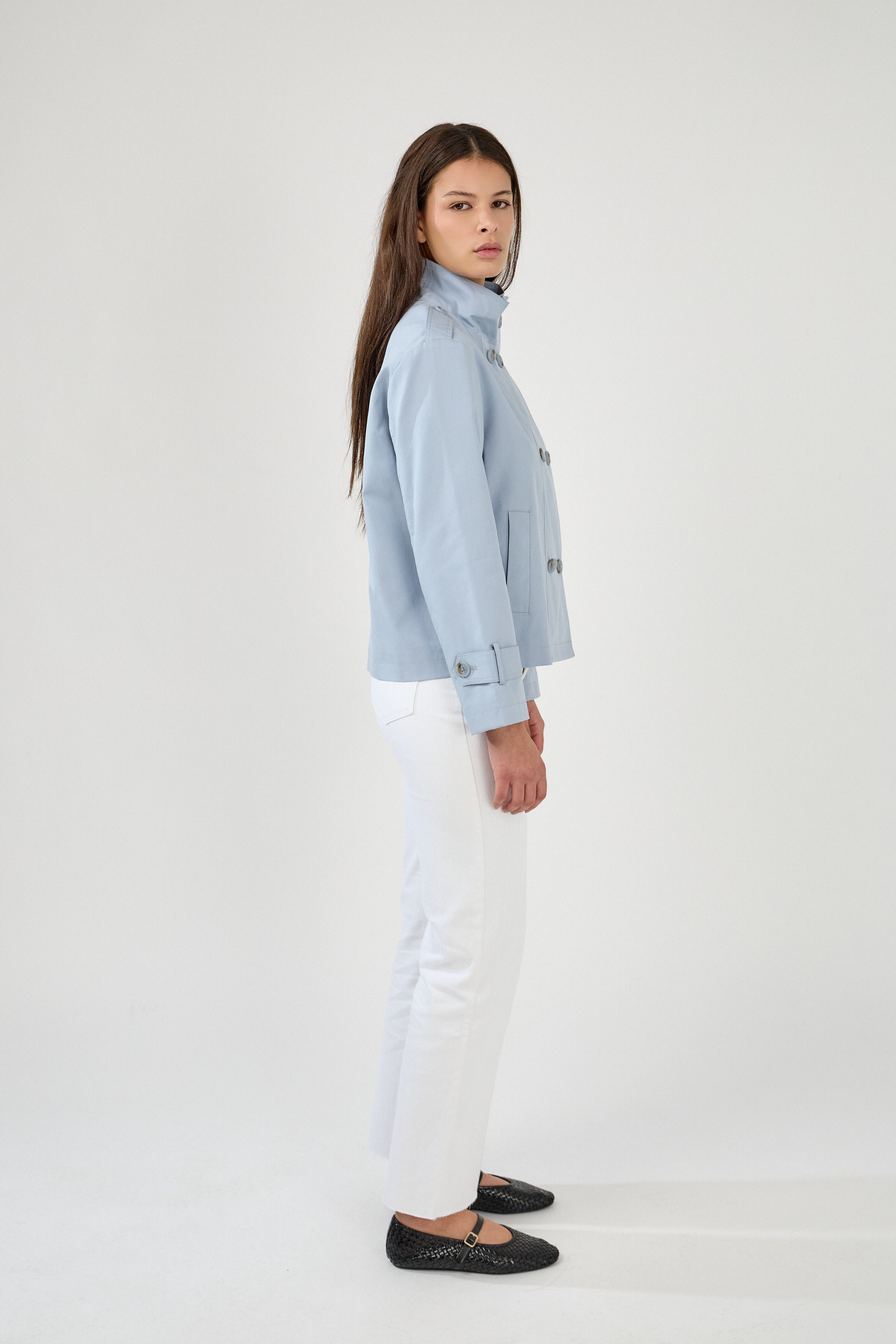 femme portant la veste trench col montant Lynara bleu ciel, tenue chic féminine, fermeture à boutons et poches raglans, pattes de serrage décorative au poignets, côté
#couleur_ciel